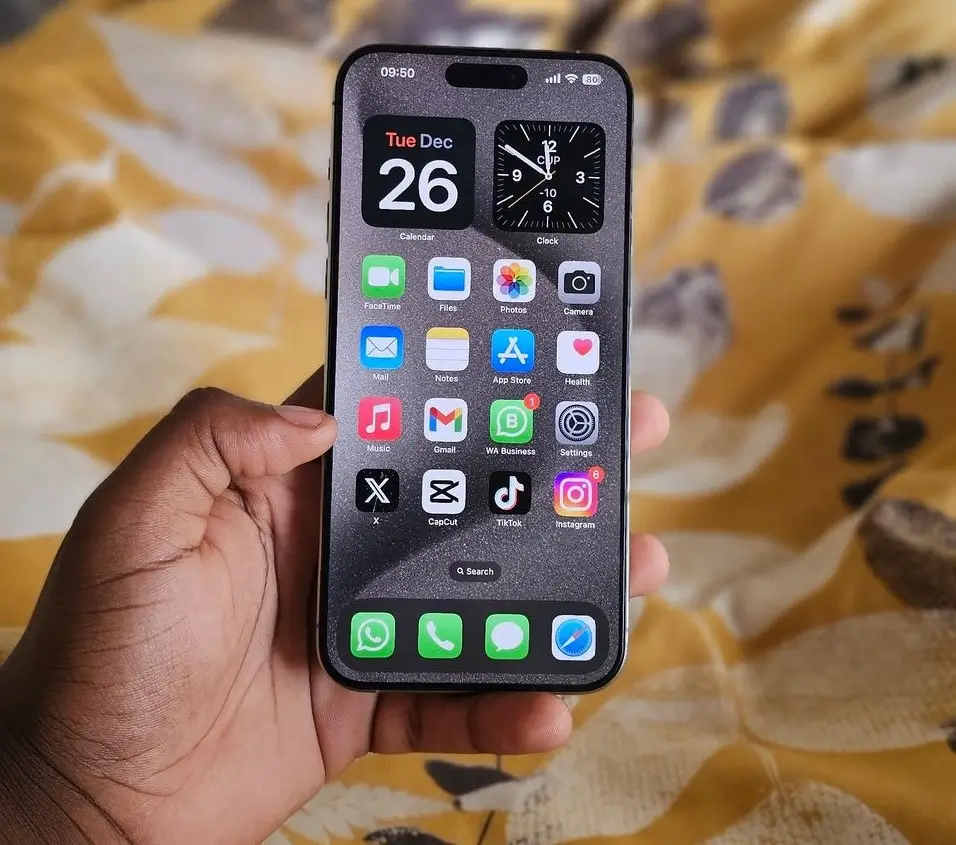 A person holding a black iPhone 15 Pro Max in their right hand, showcasing the phone's camera bump and Dynamic Island in front of a blue background.