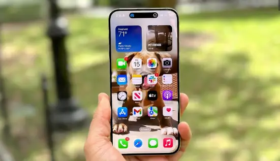 Hand holding iPhone 15 Pro Max with various app icons displayed on the home screen, set against an outdoor greenery backdrop