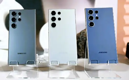 Three Samsung S24 phones in different colors with rectangular camera modules on a clear stand against a blurred background.